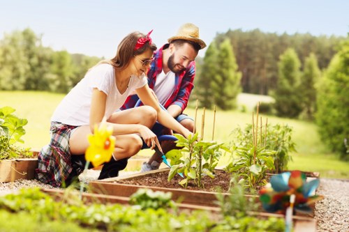 In a well-maintained garden, a woman and a man are tending to vegetable plants in raised beds filled with rich, dark soil. The woman, wearing a white t-shirt, plaid shorts, and a pink headband, is using a small gardening tool to adjust or plant in the bed, while the man, dressed in a straw hat, a red and blue plaid shirt, and dark pants, is nearby, observing or assisting. The garden features neatly bordered beds with some young vegetable plants visible, and a small decorative yellow figurine is placed among the greenery. In the foreground, there are additional garden features including a blue watering can and small garden tools, suggesting ongoing gardening activities. Behind them, the outdoor space extends into a lush, green landscape with a mix of trees and a grassy field under a bright, partly cloudy sky, creating a peaceful and productive outdoor environment typical of Fulham's suburban gardens and landscaping spaces, with natural colors of green, earthy soil, and garden tools enhancing the scene. This setting illustrates active outdoor gardening, supported by professional landscaping and garden maintenance services in the Fulham area aimed at creating vibrant, functional outdoor spaces.