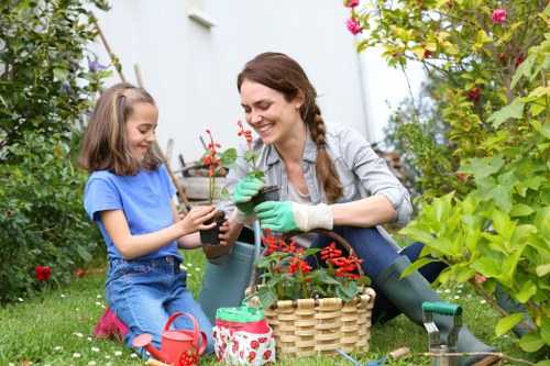 A woman and a young girl are engaging in gardening activities in a lush, well-maintained backyard garden with vibrant greenery and flowering plants. The woman is smiling while holding a small potted plant with bright red flowers, wearing gardening gloves and casual outdoor clothing. The girl, sitting on the grass, is also wearing casual clothes and pink boots, holding a small container. Surrounding them are various garden tools, a wicker basket, and watering cans, indicating gardening work. The background features a white house with potted plants and tall bushes, creating a lively and inviting outdoor space suitable for family gardening. The scene is well-lit with natural daylight, emphasizing the fresh, natural tones of the garden, consistent with professional outdoor gardening and landscaping practices near Fulham, in an environment that highlights garden nurturing and planting activity.
