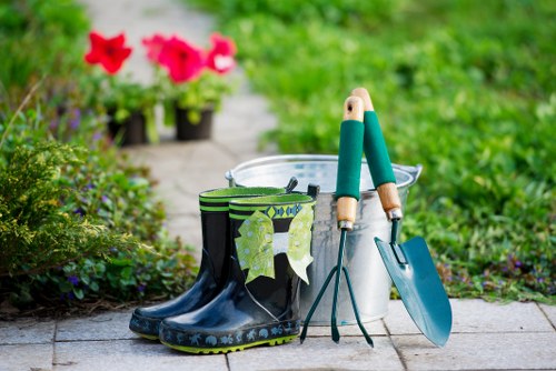 An outdoor garden scene featuring a concrete pathway with a pair of black rubber gardening boots with green and white patterned socks stuffed inside, placed beside a silver watering can, a small hand trowel with a green handle, and a pair of metal gardening shears with wooden handles. In the background, there are potted pink petunias and lush green foliage, indicating a well-maintained garden area typical of residential landscaping in Fulham. The setting suggests active gardening or outdoor maintenance, with natural daylight illuminating the scene, highlighting the vibrant colours of the flowers and greenery, and supporting local gardening and landscaping services like those offered by Landscapers Fulham.