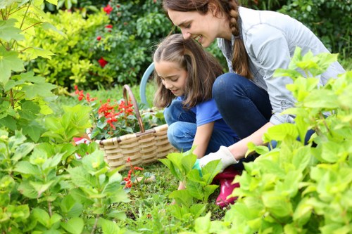 A woman and two young girls gardening in a lush outdoor space, with the woman kneeling beside a flower bed, smiling as she assists the girls who are gathering red flowers into a wicker basket. The garden features vibrant green foliage, including dense bushes, hedges, and a variety of flowering plants. The lawn area in the foreground is well-maintained with evenly cut grass, while the background displays more garden plants and a hint of potted greenery. The scene is set on a bright, mild day, with natural sunlight illuminating the garden's rich colours and textures, highlighting the natural outdoor environment typical of Fulham’s residential gardens. This image reflects professional gardening and outdoor maintenance services offered by Landscapers Fulham, focusing on garden cultivation, planting, and lawn care services to enhance outdoor spaces.