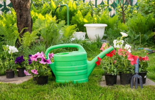 A garden scene in Fulham featuring a well-maintained grassy lawn bordered by vibrant flower beds with blooming pink, purple, and white flowers. A bright green plastic watering can is placed on the grass, surrounded by potted plants, and a small garden fork with a red handle rests nearby. In the background, there are lush green shrubs and trees, along with a white garden urn adding decorative detail. The outdoor space appears to be part of a landscaped backyard, with natural sunlight illuminating the area, highlighting the varied textures of grass, plant leaves, and flower petals, suggesting a well-cared-for garden suitable for domestic gardening services by Landscapers Fulham.
