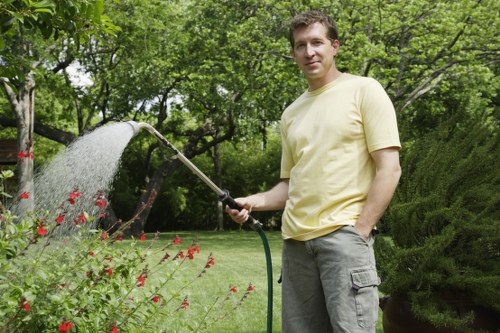 A man standing in a lush garden during daylight, watering a flower bed with a garden hose. The garden features a well-maintained lawn with dense, vibrant green grass, bordered by flowering plants with red blossoms. Behind him, there are mature trees with leafy branches providing shade, and neatly trimmed bushes and shrubs creating a structured landscape. The background includes a mix of taller trees and natural greenery, suggesting a landscaped outdoor space typical of a residential garden in Fulham. The sky appears overcast, giving soft, natural lighting that highlights the textures of the grass, plants, and garden elements. This scene reflects professional gardening and lawn care efforts, aligning with services offered by Landscapers Fulham, who specialize in maintaining outdoor spaces for local homes.