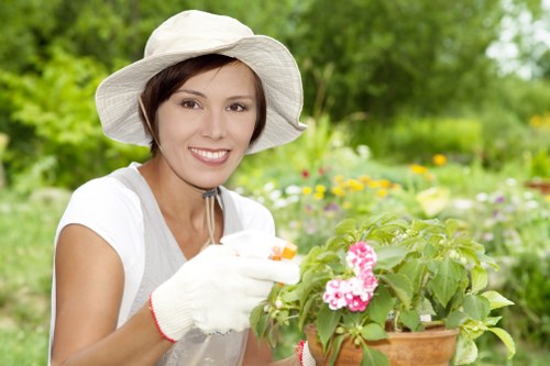 The image shows a woman in a white sun hat and gardening gloves smiling as she tends to a potted plant with pink and white flowers, set against a lush, green outdoor garden background with various plants, shrubs, and trees. She appears to be engaged in gardening activities, possibly watering or planting, in a well-maintained garden area that includes a mix of grass, flower beds, and natural foliage, typical of residential outdoor landscapes in Fulham. The lighting suggests a bright, clear day with natural sunlight illuminating the scene, highlighting the vibrant greenery and floral colours. This setting reflects professional gardening care and outdoor maintenance work carried out by Landscapers Fulham, supporting a neat, healthy, and visually appealing garden environment.