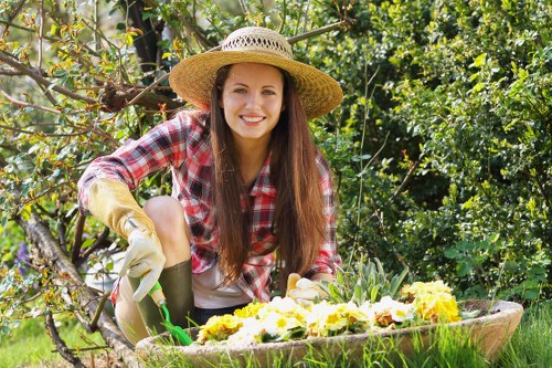 A young woman with long brown hair wearing a wide-brimmed straw hat, a red and white checkered shirt, and gardening gloves is kneeling in a lush garden, tending to a flower bed with yellow and white blossoms. The garden features a well-maintained lawn with dense green foliage, including trees and shrubs in the background, and a wooden border separating the flower beds from the grassy area. The scene is brightly lit by natural sunlight, suggesting a clear day with mild weather. This outdoor space appears to be part of a residential garden, typical of Fulham, with various plantings and a focus on cultivating colourful flowers. The woman’s focused engagement with her gardening activities reflects care for outdoor maintenance and landscaping, aligning with professional garden services offered by Landscapers Fulham.