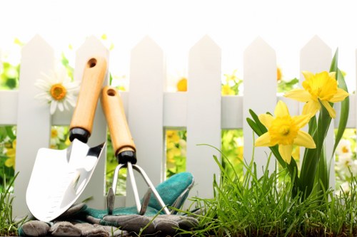 The image showcases a small garden area with a white picket fence in the background, providing a cheerful and well-maintained outdoor space. In the foreground, there is a patch of vibrant green grass with a lush, dense texture, bordered by a flower bed containing bright yellow daffodils in full bloom, along with a few white daisies. Adjacent to the flower bed, a section of soil appears freshly cultivated or prepared for planting. Resting on the soil are gardening tools, including a small hand trowel with a wooden handle, a Japanese-style hand fork with a metal head, and a pair of gardening gloves, indicating recent or ongoing garden maintenance. The lighting suggests a natural, sunny day, contributing to the vivid colours and healthy appearance of the plants. This outdoor scene exemplifies typical elements of a residential garden requiring regular lawn care and nurturing, which a professional gardening service like Landscapers Fulham can assist with, enhancing the overall appearance and health of the outdoor space in Fulham postcode area.