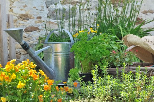 A vibrant garden scene featuring a variety of plants and garden features. In the foreground, there are bright yellow flowering plants with dense green foliage, alongside other smaller green plants with delicate leaves. A classic silver metal watering can is positioned among the plants, partially obscured by foliage. In the background, tall grass-like plants grow along a rustic stone wall, adding height and texture to the outdoor space. To the right, there is a woven straw hat resting on a container, indicating outdoor gardening activity. The garden surface appears to be composed of rich soil and possibly some paving elements, with a natural and well-maintained appearance. The overall setting suggests a peaceful, cultivated outdoor area suitable for gardening and landscaping maintenance, with natural lighting indicating a clear, possibly sunny day in Fulham or nearby within southwest London, as referenced by the local domain. This scene exemplifies typical features of a residential garden in an urban setting, maintained by professional gardeners or landscapers such as Landscapers Fulham, who provide comprehensive outdoor gardening services in the area.