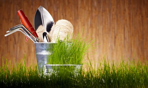 A close-up of a small garden scene featuring a patch of lush, green grass in the foreground. Behind the grass, there are two small metallic containers holding various gardening tools and supplies. One container contains a collection of hand tools such as a rake, trowel, and pruners, all with wooden or metal handles, while the other holds a ball of twine and some fresh, green grass or young plants. The background showcases a wooden surface, likely a garden shed or fence, with warm brown tones that contrast with the bright green of the garden plants. This outdoor space appears well-maintained and is typical of a suburban garden in Fulham, suitable for gardening and landscaping activities. The scene captures the preparation or maintenance phase, emphasizing readiness for outdoor gardening work in a residential setting, reinforcing the relevance of professional gardening services offered by Landscapers Fulham in the local area.