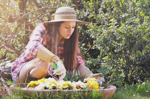 A young woman wearing a straw sun hat and a plaid shirt is gardening outdoors in a lush, well-maintained garden in Fulham. She is kneeling on the grass, wearing gardening gloves, and tending to a flower bed that features vibrant yellow and white flowers. The garden includes neatly trimmed hedges and shrubs in the background, with a mixture of green foliage and blooming plants under natural daylight, indicating a mild weather day. The scene showcases a landscaped area with a lawn patch in the foreground, surrounded by various plant borders, highlighting a typical residential garden in Fulham. This setting is suitable for professional gardening and landscaping services provided by Landscapers Fulham, emphasizing quality outdoor maintenance and garden enhancement across the local area.
