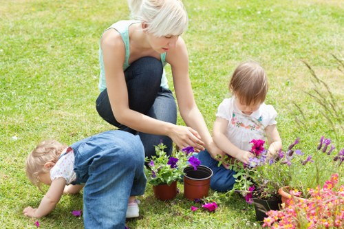 A woman with short blonde hair, dressed in a light blue sleeveless top and dark jeans, is crouched on a well-maintained, lush green lawn in a residential garden. She is engaged in planting or tending to potted flowers, with her hands near the soil. Two young children, a girl with light brown hair wearing a white dress with a floral pattern and another girl with curly blonde hair in a white top, are also involved; one is kneeling close to the ground, examining the flowers, while the other is sitting with a small bouquet of pink flowers. The garden features a vibrant border of flowering plants and bushes with purple and pink blooms. The scene is outdoors on a clear day, with soft natural lighting accentuating the varied textures of the grass, soil, and flowers. The background reveals additional garden elements, such as a paved path or patio, typical of Fulham gardens. This outdoor space appears to be well cared for, reflecting professional gardening services. The overall setting is cheerful and tranquil, emphasizing gardening activities suitable for a family garden environment in London.