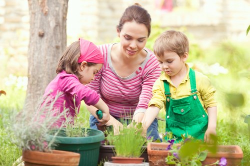 A woman and two children are gardening outdoors in a well-maintained backyard with a lush green lawn and mature trees. The woman, dressed in a pink-and-white striped top, is smiling as she assists the children with planting or tending to potted plants arranged on a wooden table. One child, wearing a pink headscarf and pink top, is actively handling a small gardening tool and focusing on a plant in a green pot. The other child, in a yellow shirt with green overalls, observes the activity, holding a small gardening tool. The garden features a combination of flower pots with blooming plants, soil beds, and a wooden decking area. Bright natural light indicates a clear day, with gentle sunshine illuminating the scene. In the background, the garden's natural environment includes a stone wall and leafy foliage, creating a peaceful outdoor space. This scene reflects a family-friendly gardening activity, suitable for local outdoor maintenance and landscaping services in Fulham, helping to create inviting garden environments such as this one.