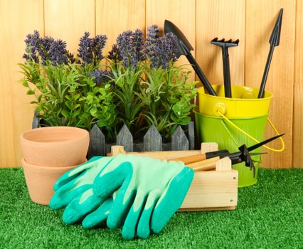 The image displays a tidy gardening storage scene on a lush green artificial turf surface, set against a wooden fence backdrop. In the foreground, a pair of bright turquoise gardening gloves rests on a wooden tool tray, alongside several small terracotta plant pots stacked one inside the other. To the right, a yellow plastic bucket contains garden tools such as a trowel, a hand rake, and a cultivator, all with black handles. Behind the bucket, a black watering can and a long-handled garden fork are visible, leaning against the fence. In the background, there are small potted lavender plants with purple flowers, arranged in a neat row, emphasizing practical garden planting and maintenance. The scene suggests an organized outdoor space prepared for planting or gardening activities, suitable for a residential garden in Fulham or nearby London areas, managed by Landscapers Fulham as part of their gardening services. The weather appears clear, with natural light illuminating the scene effectively, highlighting the vibrant colors and textures of the gardening tools and plants.