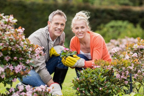A man and a woman are gardening in a lush outdoor space, kneeling among blooming pink and white flowers, with the man wearing grey and the woman in an orange jacket. They are smiling at the camera and wearing yellow gloves, appearing to be planting or tending to the garden. The garden features well-maintained flower beds, dense green foliage, and small shrubs. In the background, there are trees and a slightly blurred outdoor environment, suggesting a serene, landscaped backyard or front garden. The scene is set in natural daylight, with a clear sky providing bright and even lighting, highlighting the vibrant colours of the flowers and the greenery. This outdoor setting reflects professional gardening and landscaping work commonly undertaken by firms such as Landscapers Fulham, servicing residential gardens in Fulham and surrounding areas, with lush, well-kept plantings and a carefully maintained lawn area visible in the image.